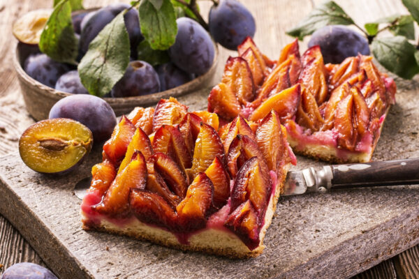 Zwei Zwetschkenkuchen-Stücke auf einem Holzbrett umgeben von frischen Zwetschken